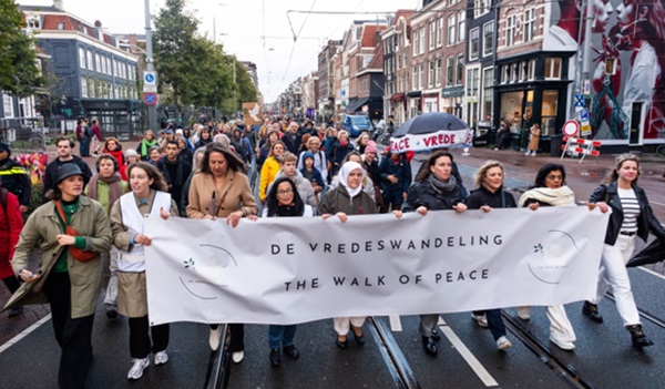 Een groep mensen die door de stad heen lopen met een spandoek, waarop staat: De vredeswandeling, The walk of peace